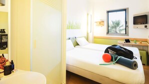 Habitación de hotel con cama, escritorio con computadora, televisor y ventana con vista a palmeras.