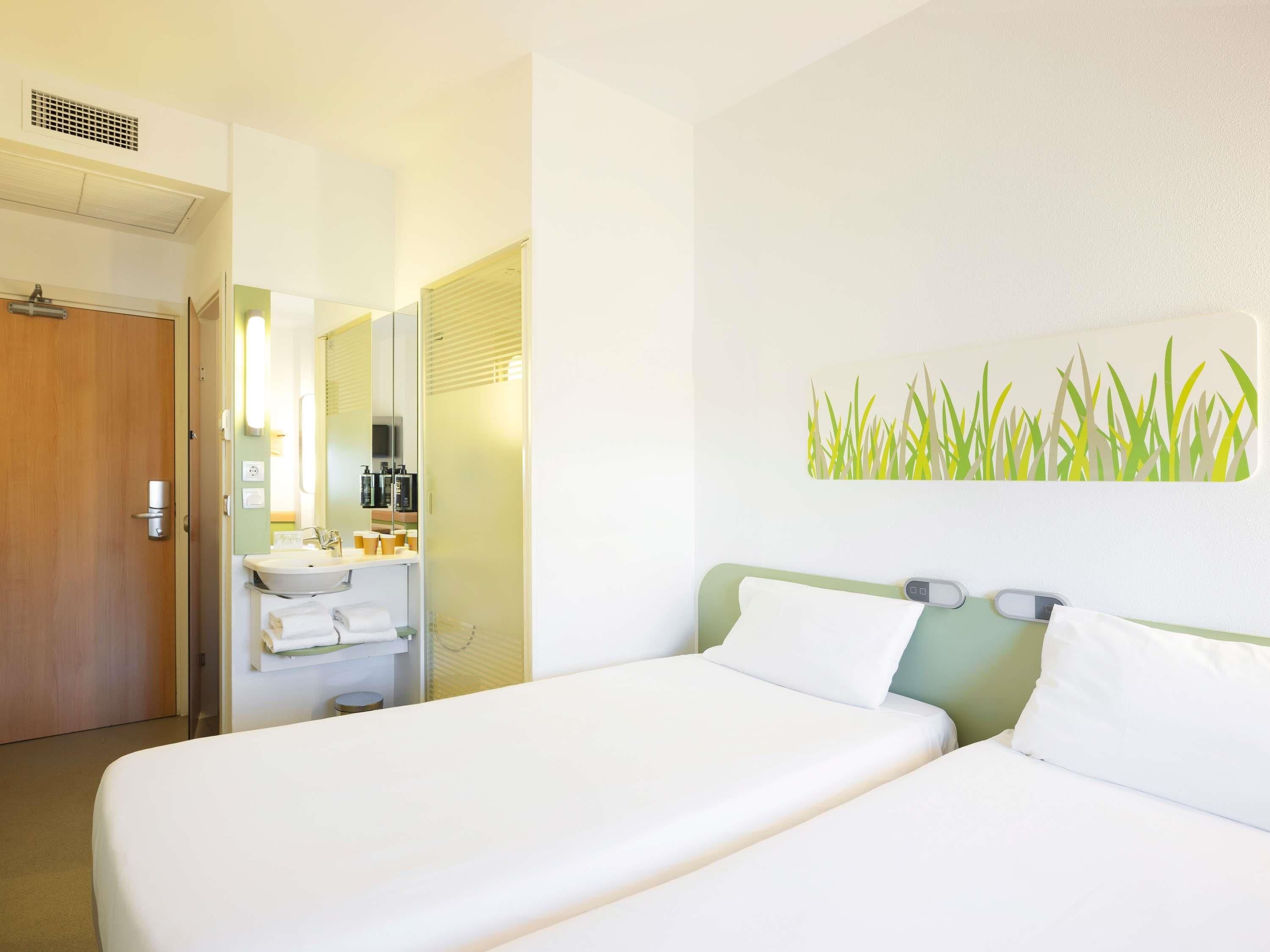 A hotel room with a bed, a bathroom with a sink and mirror, and a wall with a grass painting.