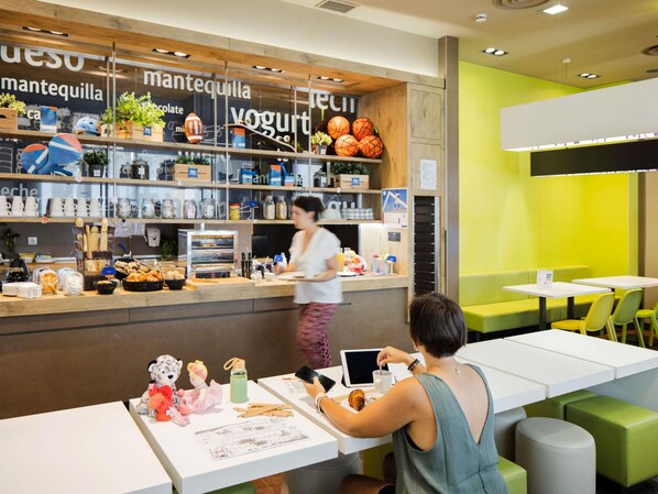 A café with a counter displaying various food items and a person serving. There are tables with chairs and a green wall in the background. - Ibis Budget Sevilla Aeropuerto (Seville)