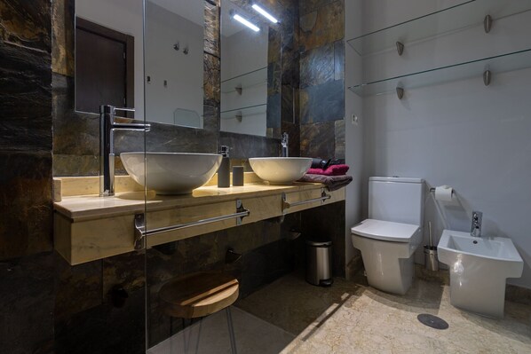 Une salle de bain moderne avec un mur en pierre, un grand miroir, un meuble-lavabo double avec des vasques sur pied, des toilettes, un bidet et une poubelle.