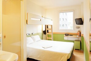 Een kleine hotelkamer met een stapelbed, een bureau met stoel, een televisie en een raam met uitzicht op gebouwen.