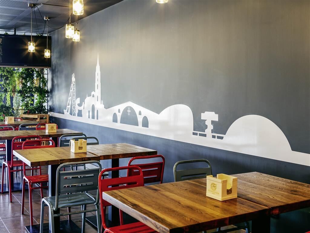Un restaurante con mesas de madera y sillas rojas, un mural de un horizonte urbano y luces colgantes.