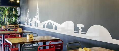 Un restaurante con mesas de madera y sillas rojas, un mural de un horizonte urbano y luces colgantes.