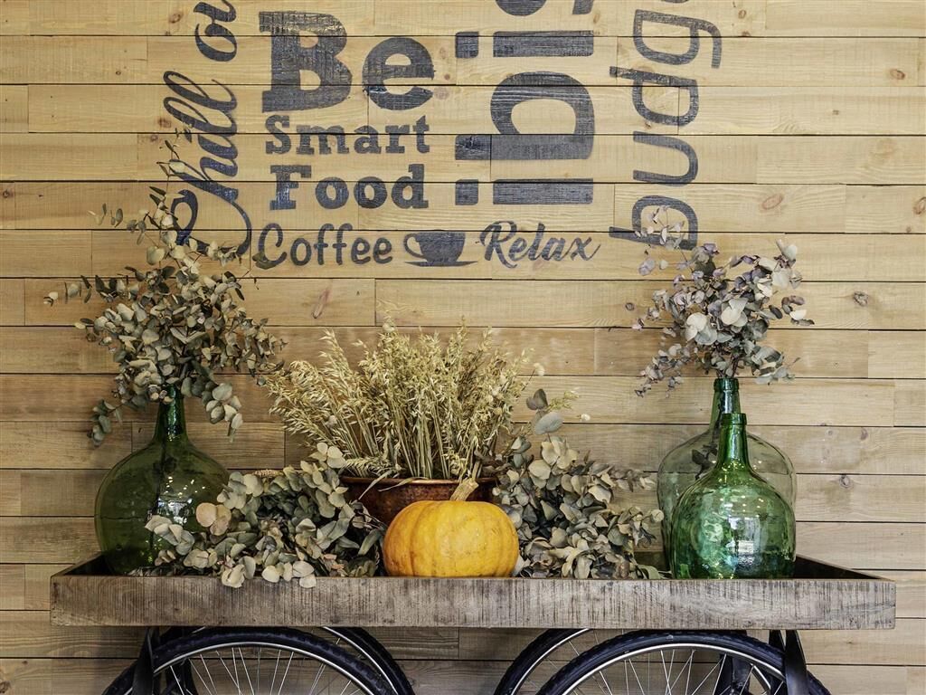 Un muro di legno con un cartello che recita “Be smart Food Coffee Relax”. Due bottiglie di vetro verde con fiori secchi e una piccola zucca su un ripiano di legno.