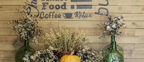 Eine Holzwand mit einem Schild, das "Be smart Food Coffee Relax" zeigt. Auf einem Holzboden stehen zwei grüne Glasflaschen mit getrockneten Blumen und ein kleiner Kürbis.