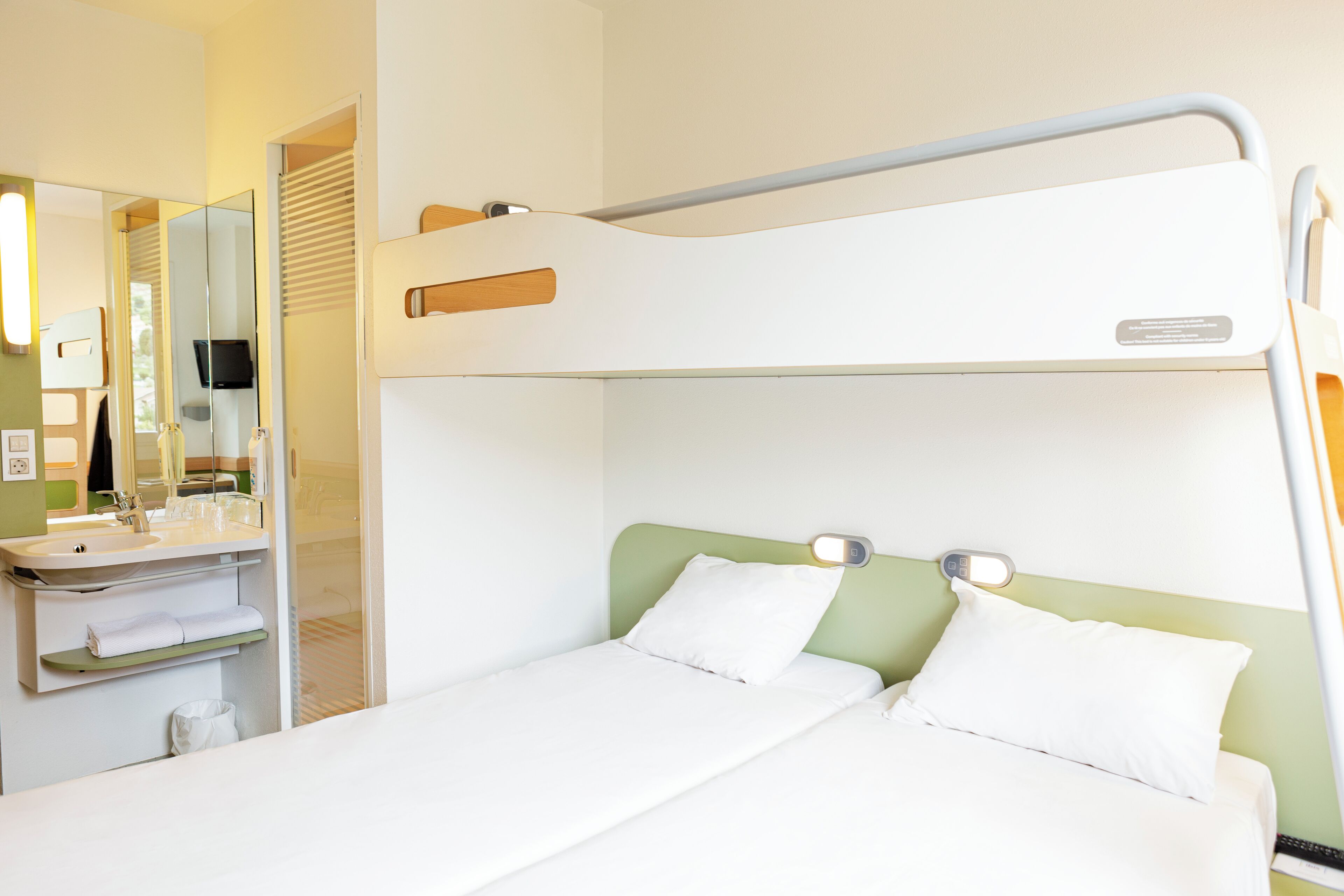 Een stapelbed met een groen hoofdeinde, een nachtkastje met een spiegel, en een wastafel in een hotelkamer.