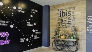 A lobby with a black wall featuring a map of San Francisco landmarks, a wooden wall with a word cloud of travel-related words, and a cart with flowers and bottles. - ibis budget Oviedo (Oviedo)
