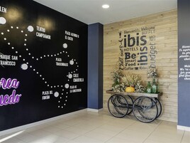 Un vestíbulo con una pared negra que muestra un mapa de lugares emblemáticos de San Francisco, una pared de madera con una nube de palabras relacionadas con viajes, y un carrito con flores y botellas.