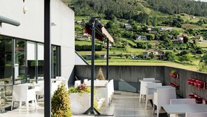 A terrace with white tables and chairs, a red awning, and a view of a green hillside with houses. - ibis budget Oviedo (Oviedo)