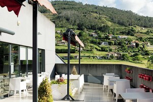 Una terraza con mesas y sillas blancas, un toldo rojo y vistas a una ladera verde con casas.