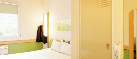 A hotel room with a bed, a window, a coat rack, a wall-mounted light, and a mirror.