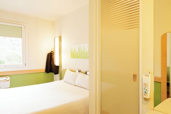 Una habitación de hotel con cama, ventana, perchero, luz empotrada y espejo.