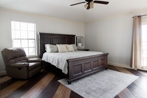 Un dormitorio con una cama de madera oscura, un sillón reclinable, un ventanal con persianas, un ventilador de techo y una alfombra en el suelo.