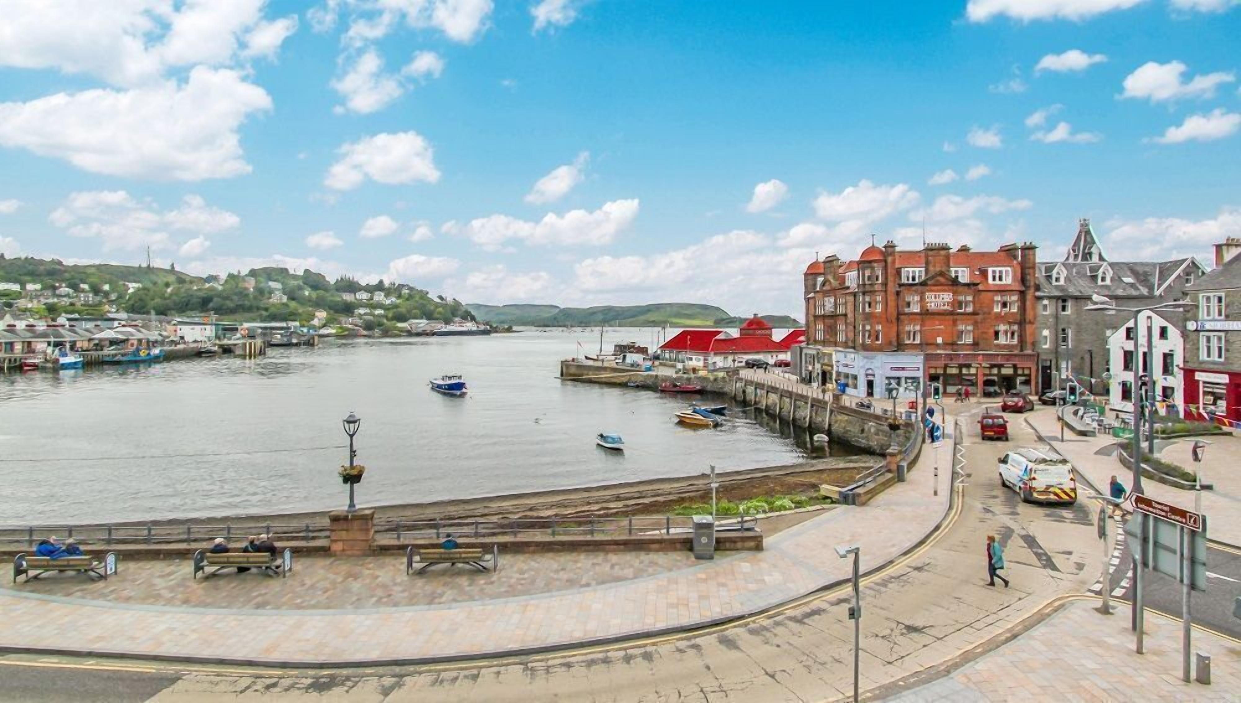 Um calçadão à beira-mar com bancos, um píer com barcos e edifícios com telhados vermelhos.