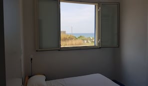A bedroom with a single bed, a window with a view of the sea, and a wall-mounted light fixture.