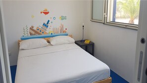 A bed with a white mattress and pillows, a nightstand with a lamp, and a wall mural of a shipwreck.