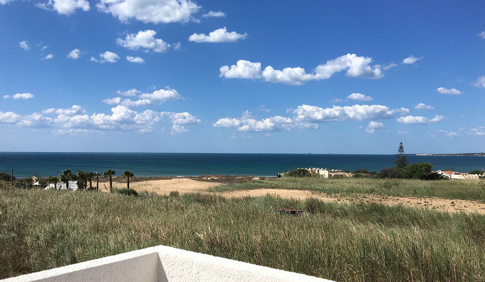 A coastal landscape with a sandy beach, grassy dunes, and a clear blue sky with scattered clouds.