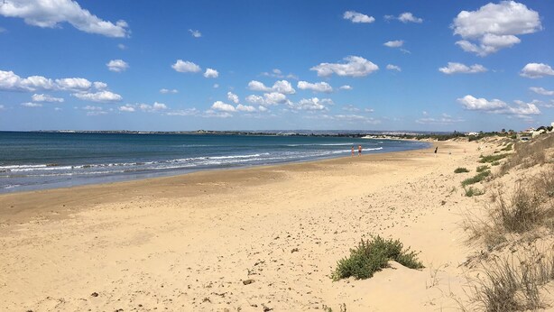 Una amplia playa de arena con olas suaves, cielo azul despejado y nubes dispersas.