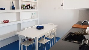 A modern kitchen with white cabinetry, a blue countertop, and a white dining table with four chairs. There are wine bottles on the upper shelf and a sink with a faucet.