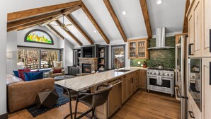 A modern kitchen with a large island, wooden beams, and a stone fireplace. - Creekside Hideaway (Crested Butte)