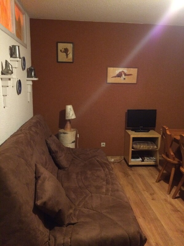 A living room with a brown sofa, a television on a wooden stand, a lamp, and a small table.