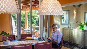 Ein Mann arbeitet an einem Schreibtisch in einem hellen Raum mit großen Fenstern, einer Küchenzeile und einem Essbereich mit roten Stühlen und einem Tisch.
