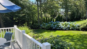 A wooden deck with white railings and outdoor furniture, surrounded by lush greenery and flowering plants. - Last Chance for August — A Bright, Breezy Stay on the Right Side of the Bridge (Harwich)