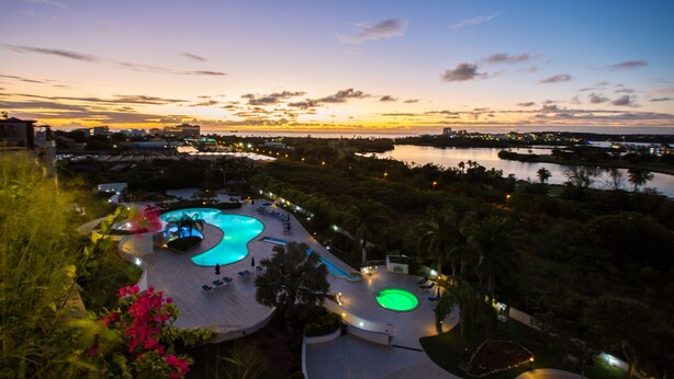 A resort with a large swimming pool, palm trees, and a view of the city at sunset. - Modern Penthouse with amazing view- Private Pool (Cupecoy)