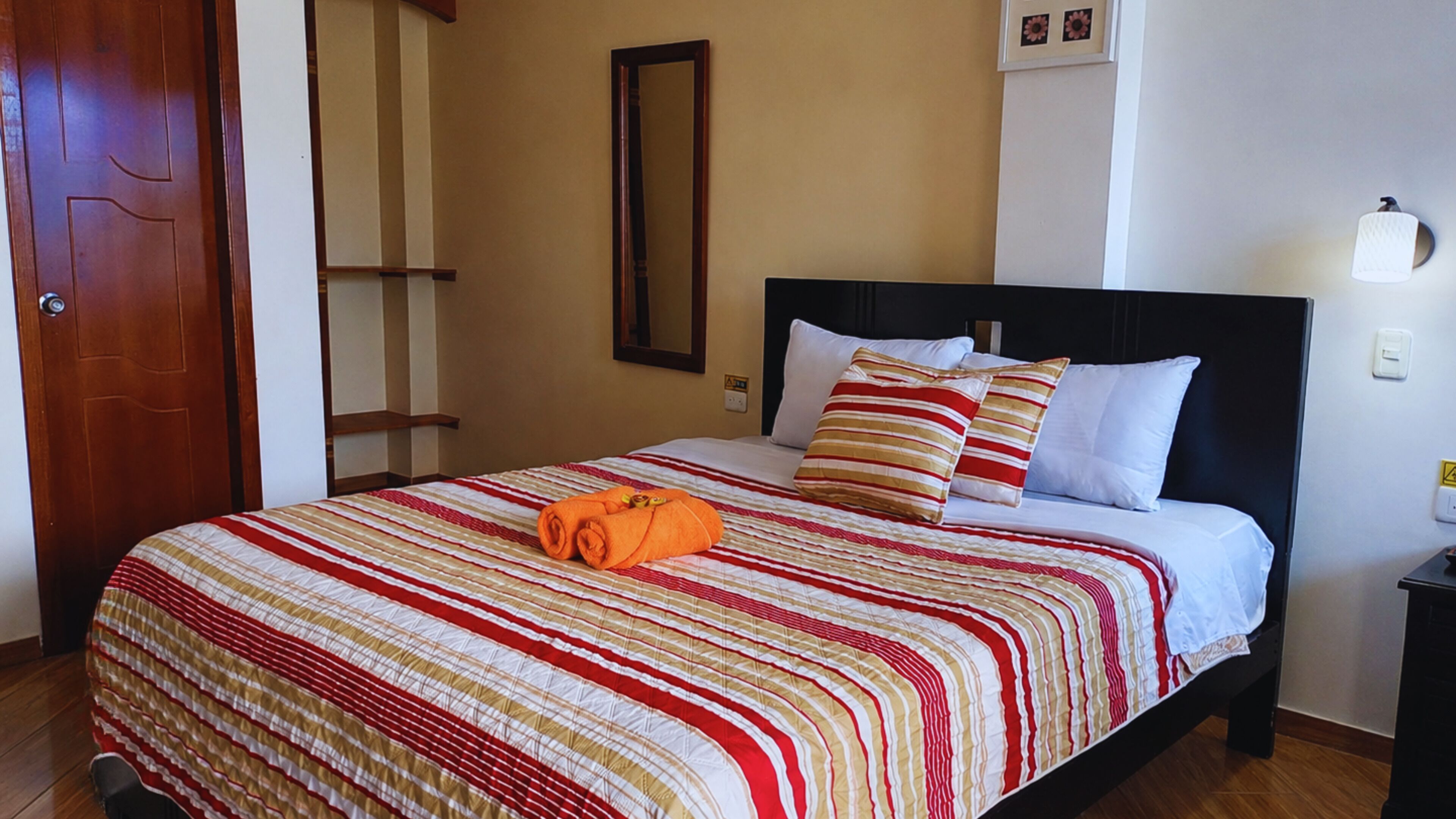 A hotel room with a bed, a wooden door, a mirror, a nightstand, and a wall-mounted light fixture.