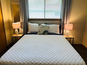 A hotel room with a large bed, two bedside lamps, a window with blinds, and a mirror on the wall. -  Chicagoland/Northwest Indiana Corporate Condo Travel Nurses /Oil Refinery (Merrillville)