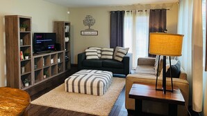 Ein Wohnzimmer mit Sofa, Hocker, Fernseher, Bücherregal und einer Lampe.