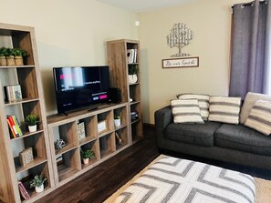 A living room with a flat-screen TV, wooden shelving with books and plants, a grey sofa with striped cushions, and a 'Be our Guest' sign on the wall.