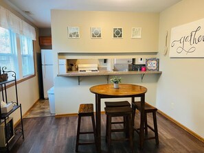Cocina compacta con comedor, que incluye una mesa y sillas de madera, una pequeña planta en maceta y una estantería con utensilios de cocina.