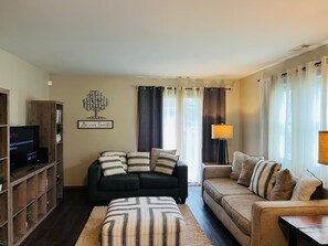 A living room with a dark-colored sofa, a striped ottoman, a wooden bookshelf, a flat-screen TV, and a wall-mounted lamp. -  Chicagoland/Northwest Indiana Corporate Condo Travel Nurses /Oil Refinery (Merrillville)