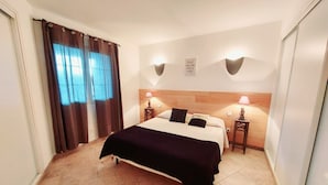 A hotel room with a bed, two bedside tables, a headboard, a window with curtains, and a wall-mounted light fixture. - Apartments in Residence with swimming pool (Sainte-Lucie de Porto-Vecchio)