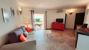 A living room with a sofa, a TV on a red cabinet, and a dining area with a table and chairs. - Apartments in Residence with swimming pool (Sainte-Lucie de Porto-Vecchio)