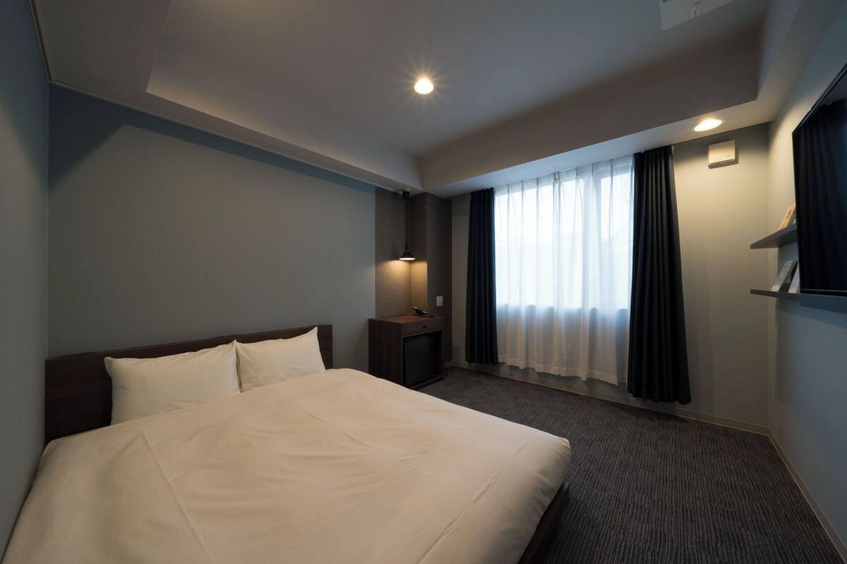 Habitación de hotel con una cama grande, una mesita de noche con lámpara, un televisor y una ventana con cortinas.