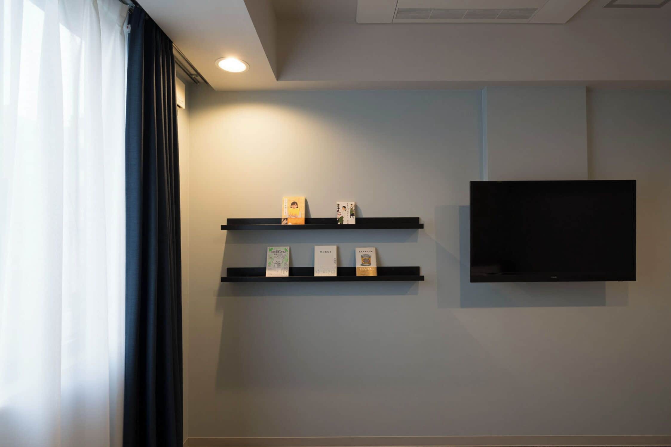 A room with a flat-screen TV mounted on the wall, three shelves with items, and a window with curtains.