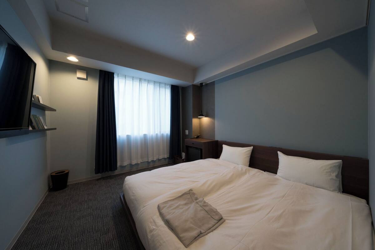 Una habitación de hotel con una cama grande, una mesita de noche con lámpara, un cesto de basura pequeño y una ventana con cortinas.