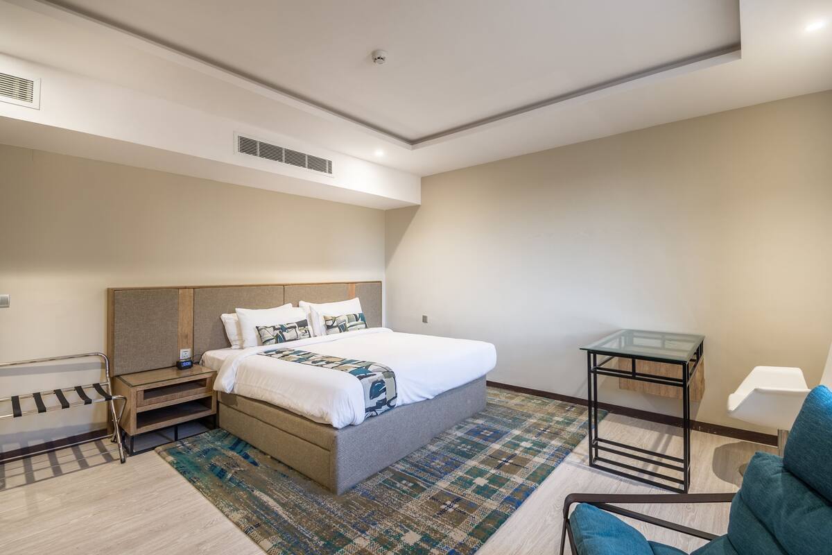 A modern hotel room with a large bed, a bedside table, a chair, and a small table with a lamp.