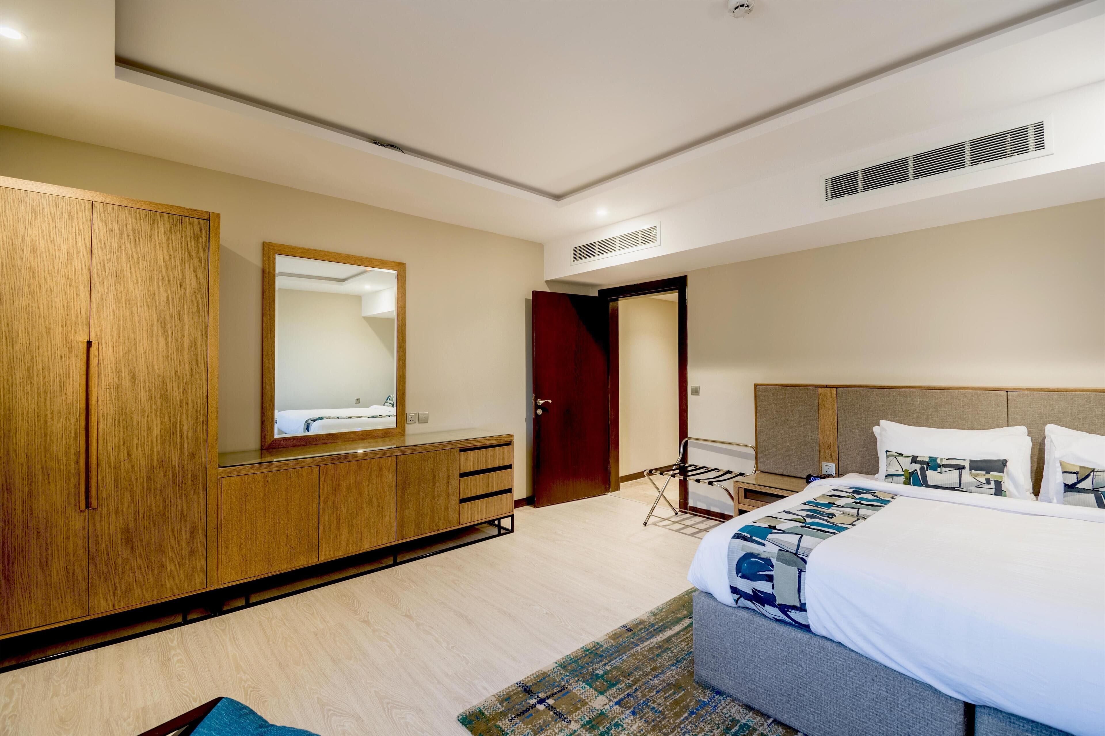 A modern hotel room with a large bed, a wardrobe, a mirror, and a door leading to another room.