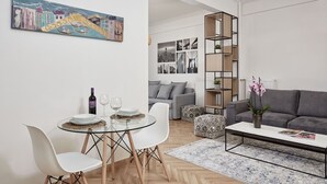 A modern living room with a glass dining table, white chairs, a grey sofa, and a coffee table. There is a bookshelf with plants and a cityscape artwork on the wall. - Kolonaki Square Executive Apartment (Athens)