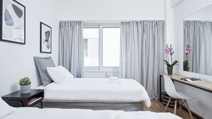 A modern hotel room with a bed, a desk, and a chair. There are two windows with curtains, a potted plant, and framed artwork on the wall. - Kolonaki Square Executive Apartment (Athens)