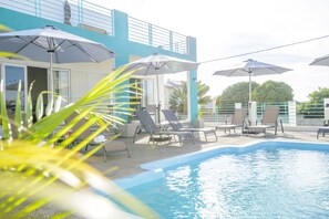 A pool area with lounge chairs and umbrellas, a building with a blue exterior, and a clear sky.