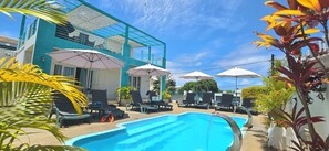 A swimming pool area with lounge chairs and umbrellas, a building with balconies, and tropical plants.