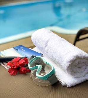 A poolside setting with a white towel, a pair of swimming goggles, a red flower, and a blue booklet on a woven chair.