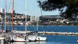 A marina with several sailboats docked, a rocky shoreline, and a cityscape in the background.