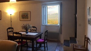 A dining area with a wooden table, chairs, a vase with dried flowers, a framed picture on the wall, and a window with a view of mountains. - Stay in a 16th century three story home in the Provence countryside. (Mison)