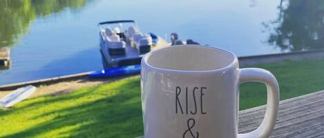 Une tasse blanche sur laquelle on peut lire les mots « RISE & SHINE », posée sur une terrasse en bois donnant sur un lac paisible, avec un bateau visible au loin.