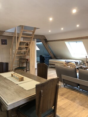 A loft-style room with a wooden dining table, chairs, a staircase, a bed with a striped blanket, and a skylight.